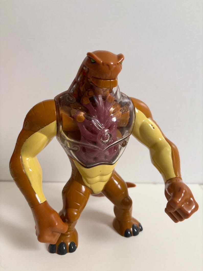 May include: A brown and yellow action figure of a reptilian humanoid alien with a clear chest and a red-purple center. The figure has a muscular build, with a tail and black claws. The figure is standing on a white surface.