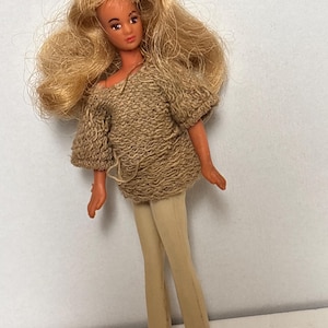 May include: A vintage doll with long blonde hair, wearing a tan knit top and matching pants. The doll has a painted face with red lips and is posed standing up. The doll's outfit is a retro style.