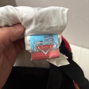 May include: A fabric tag attached to a textile item featuring the Disney Pixar Cars logo in red and white text against a blue background. The tag is sewn into a white fabric seam and includes additional white care labels partially visible underneath.