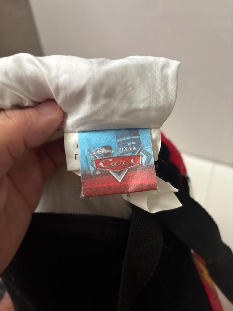 May include: A fabric tag attached to a textile item featuring the Disney Pixar Cars logo in red and white text against a blue background. The tag is sewn into a white fabric seam and includes additional white care labels partially visible underneath.
