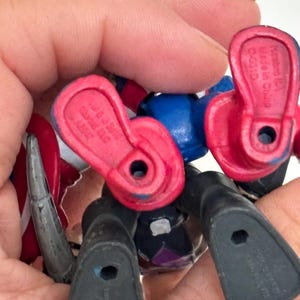 May include: A close-up of several small action figure feet and legs. The figures are primarily black, red, and blue. The feet have holes in the soles. The figures are held in a hand.