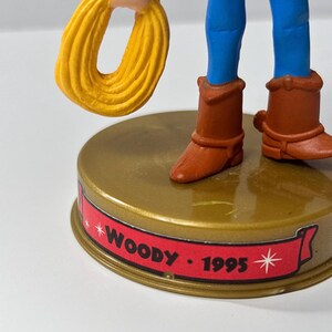 May include: A figurine of a cowboy character, featuring blue pants, brown boots, and a yellow rope. The base is gold with a red banner that reads "Woody - 1995".