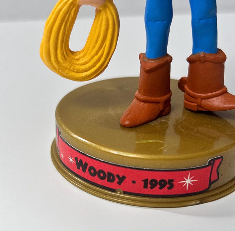 May include: A figurine of a cowboy character, featuring blue pants, brown boots, and a yellow rope. The base is gold with a red banner that reads "Woody - 1995".