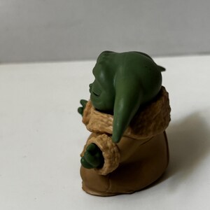 May include: A green figurine of a character with large ears, wearing a brown coat with a tan fur-like trim. The figurine is posed with its arms outstretched, and the details are clearly visible.