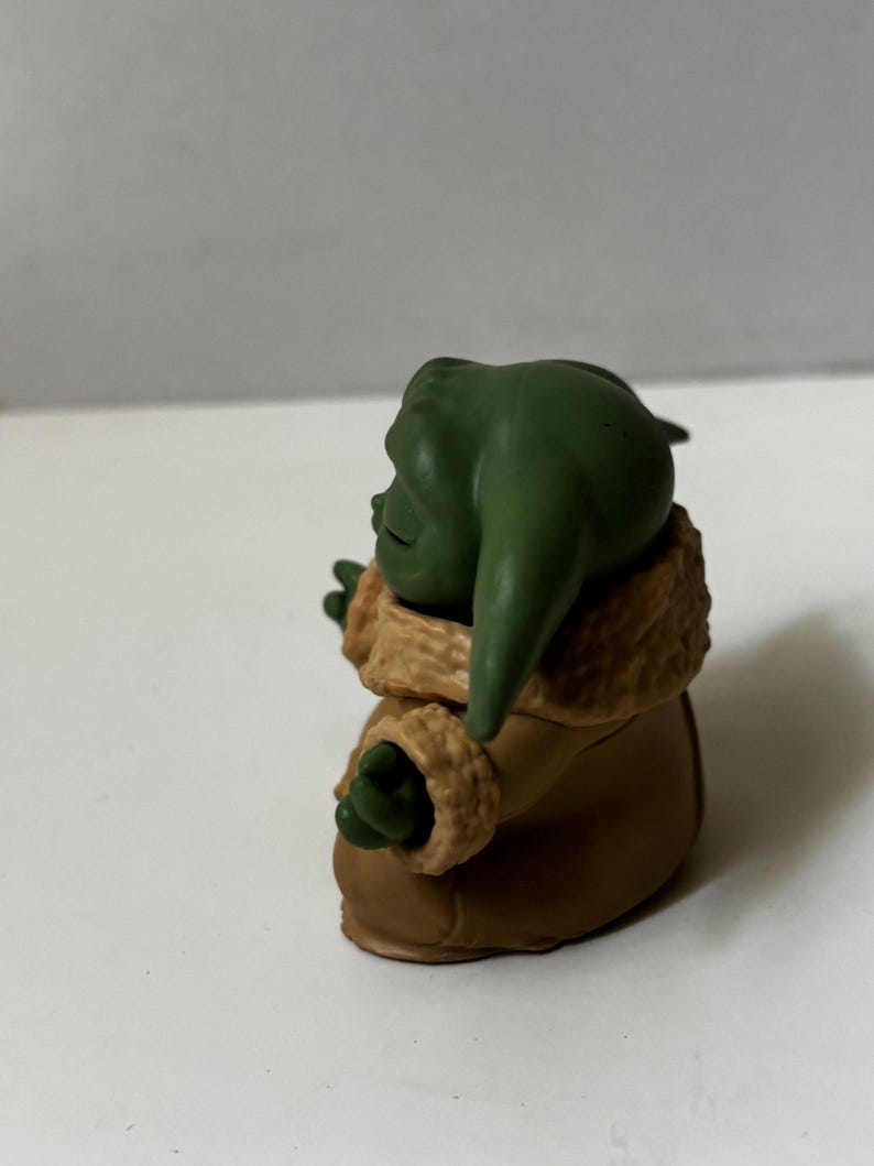 May include: A green figurine of a character with large ears, wearing a brown coat with a tan fur-like trim. The figurine is posed with its arms outstretched, and the details are clearly visible.