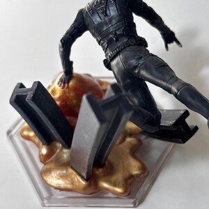 May include: A black action figure in a dynamic pose, wearing a tactical suit and helmet. The figure is positioned above a gold-colored, textured base with black geometric shapes, all set on a clear, hexagonal platform.