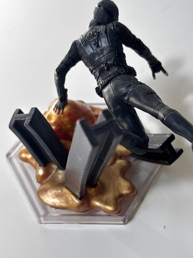 May include: A black action figure in a dynamic pose, wearing a tactical suit and helmet. The figure is positioned above a gold-colored, textured base with black geometric shapes, all set on a clear, hexagonal platform.