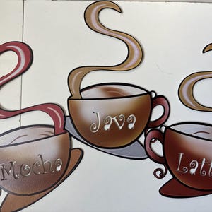 May include: Three decorative wall cutouts shaped like coffee cups, each featuring a swirling steam trail. The cups are labeled Mocha, Java, and Latte in a stylized font. The Mocha cup is reddish-brown, the Java cup is tan, and the Latte cup is dark brown.