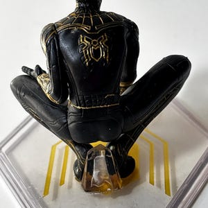 Puede incluir: Una figura de Spider-Man negra y dorada en posici&oacute;n agachada. La figura tiene detalles dorados y un emblema de ara&ntilde;a en la espalda. Se coloca sobre una base hexagonal transparente con l&iacute;neas doradas.