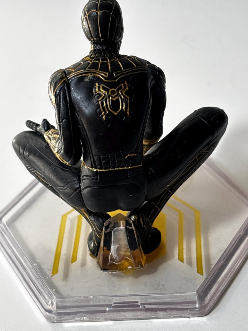 Puede incluir: Una figura de Spider-Man negra y dorada en posici&oacute;n agachada. La figura tiene detalles dorados y un emblema de ara&ntilde;a en la espalda. Se coloca sobre una base hexagonal transparente con l&iacute;neas doradas.
