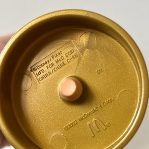 May include: A gold-colored plastic toy base with a light peach-colored peg in the center. The base has the text "Disney/Pixar MFG. FOR MCD CORP. CHINA/CHINE C-SN" and "&copy;2002 McDonald's Corp." and the McDonald's logo.