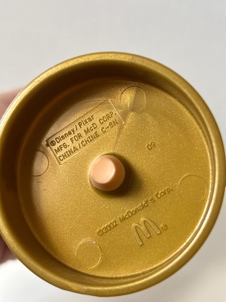 May include: A gold-colored plastic toy base with a light peach-colored peg in the center. The base has the text "Disney/Pixar MFG. FOR MCD CORP. CHINA/CHINE C-SN" and "&copy;2002 McDonald's Corp." and the McDonald's logo.