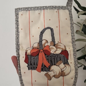 May include: Oven mitt with a mushroom basket design. The mitt features a cream background with red vertical stripes and brown polka dots. A gray basket filled with mushrooms is tied with an orange bow. The edges are trimmed with a floral pattern.