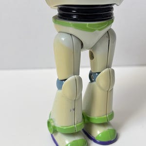 以下が含まれることがあります： バズ・ライトイヤーのアクションフィギュアの下半身のクローズアップ。フィギュアの脚はオフホワイトで、緑と紫のアクセントが付いています。ブーツには緑と紫のディテールがあります。