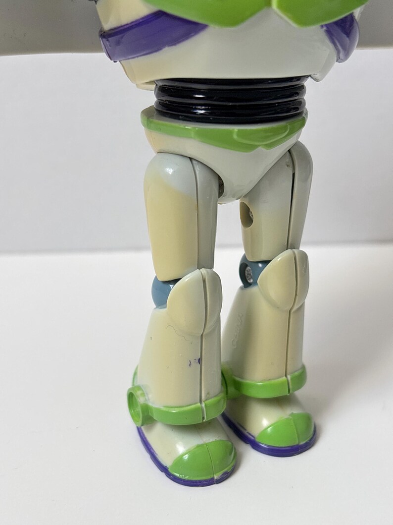 以下が含まれることがあります： バズ・ライトイヤーのアクションフィギュアの下半身のクローズアップ。フィギュアの脚はオフホワイトで、緑と紫のアクセントが付いています。ブーツには緑と紫のディテールがあります。