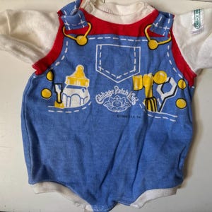 May include: A short-sleeved white and blue romper featuring a printed design of red straps, yellow buttons, a baby bottle, a hammer, and a wrench. The front displays the text Cabbage Patch Kids and 1984 OAA Inc. with a small white tag on the sleeve.