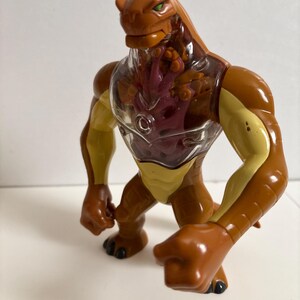 May include: A brown and yellow action figure of a reptilian alien with green eyes. The figure has a clear chest cavity revealing a red and purple interior. The toy is posed in a dynamic stance, with its fists clenched.