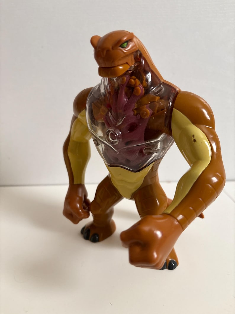 May include: A brown and yellow action figure of a reptilian alien with green eyes. The figure has a clear chest cavity revealing a red and purple interior. The toy is posed in a dynamic stance, with its fists clenched.