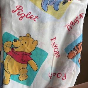 May include: A white fabric featuring a repeating pattern of Winnie the Pooh characters including Pooh, Piglet, Tigger, and Eeyore. Each character is placed within a colored diamond shape, accompanied by their name printed in a red cursive font.