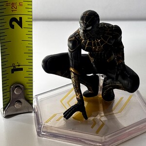 Puede incluir: Figura de Spider-Man negro y dorado en posici&oacute;n agachada, de aproximadamente 5 cm de altura, sobre una base transparente con detalles dorados. La figura tiene patrones de telara&ntilde;a detallados.