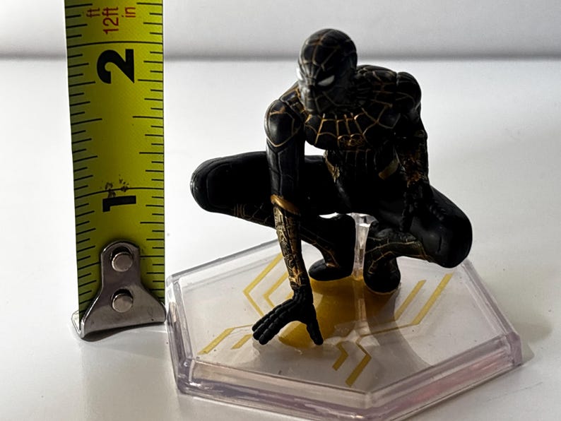 Puede incluir: Figura de Spider-Man negro y dorado en posici&oacute;n agachada, de aproximadamente 5 cm de altura, sobre una base transparente con detalles dorados. La figura tiene patrones de telara&ntilde;a detallados.