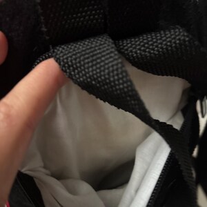 May include: A black bag with a white interior lining and black woven straps. The bag features a black zip closure and a red fabric accent visible at the top edge. The interior is made of a plain white material, and the exterior is black.