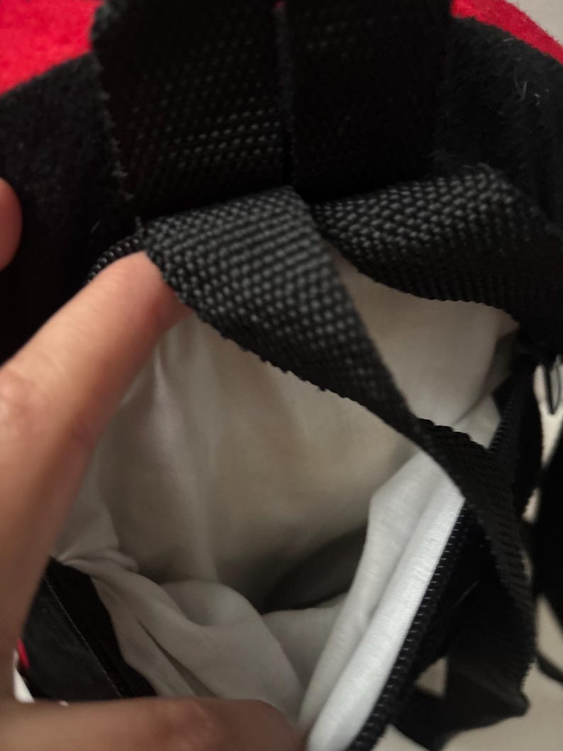 May include: A black bag with a white interior lining and black woven straps. The bag features a black zip closure and a red fabric accent visible at the top edge. The interior is made of a plain white material, and the exterior is black.