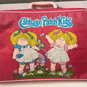 May include: A rectangular pink suitcase featuring a Cabbage Patch Kids logo and a graphic of two blonde dolls in white dresses with blue and pink accents, gardening with a small shovel and watering can amidst colorful flowers and butterflies.