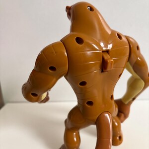 May include: A brown action figure of a creature with a long tail, muscular arms, and legs. The figure has several circular holes and a small, rectangular piece on its back. The figure is posed in a crouching position.