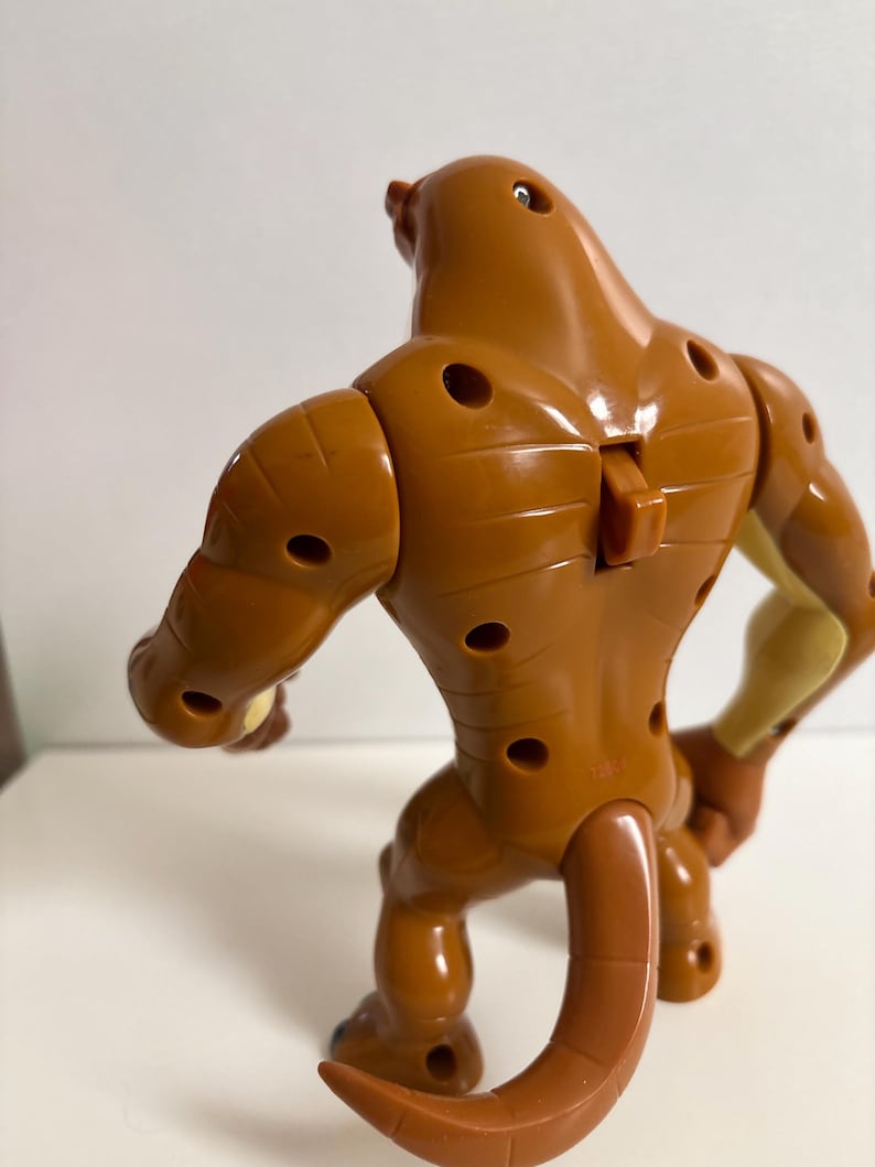 May include: A brown action figure of a creature with a long tail, muscular arms, and legs. The figure has several circular holes and a small, rectangular piece on its back. The figure is posed in a crouching position.