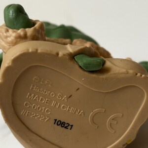 May include: A close-up of a tan-colored plastic toy figurine. The underside of the figurine is visible, with text that includes "Hasbro SA" and "Made in China". The figurine has green and tan details.
