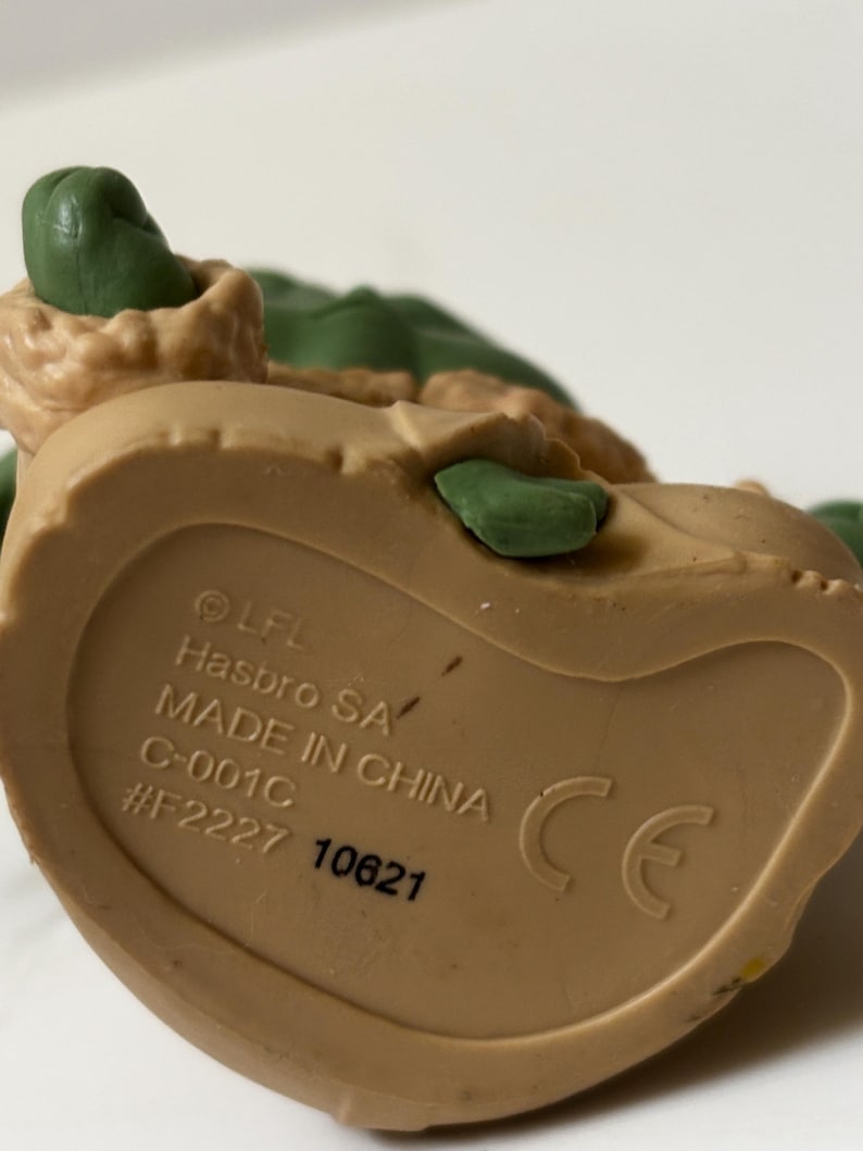 May include: A close-up of a tan-colored plastic toy figurine. The underside of the figurine is visible, with text that includes "Hasbro SA" and "Made in China". The figurine has green and tan details.
