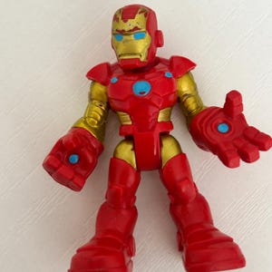 May include: A red and gold Iron Man action figure. The figure has a red body with gold accents on the arms, head, and joints. The eyes are blue. The figure is standing with its arms outstretched.
