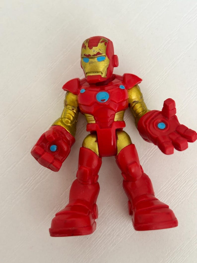 May include: A red and gold Iron Man action figure. The figure has a red body with gold accents on the arms, head, and joints. The eyes are blue. The figure is standing with its arms outstretched.