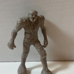 May include: A small, grey plastic monster figurine with a skeletal face and torn clothing. The figure has a belt and stands with its arms at its sides. The monster's skin appears textured, and it is on a white surface.
