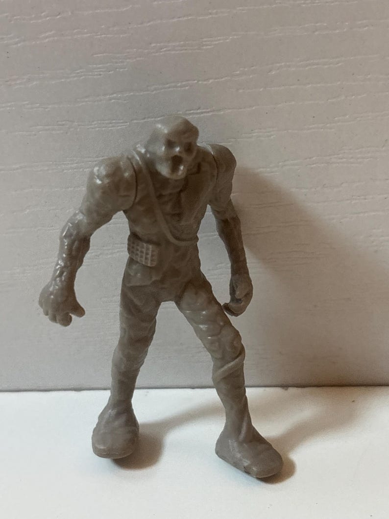 May include: A small, grey plastic monster figurine with a skeletal face and torn clothing. The figure has a belt and stands with its arms at its sides. The monster's skin appears textured, and it is on a white surface.