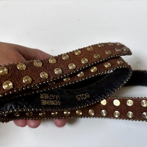 May include: Brown leather belt with gold-colored studs and rivets. The belt is marked "GENUINE LEATHER" and "ST08". The belt is a classic accessory for jeans or pants.