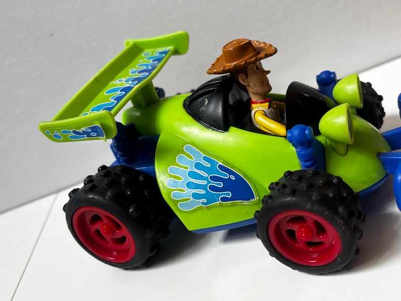 以下が含まれることがあります： 黒いタイヤと赤いリムが付いた、緑と青のおもちゃのレーシングカー。運転席にはウッディのフィギュアがいます。車には、青と白の波模様の緑色のスポイラーが付いています。