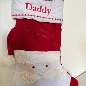 May include: Red Christmas stocking with a Santa Claus face design. The stocking has a white cuff with the word "Daddy" embroidered in red. The cuff is trimmed with red and white checkered fabric.
