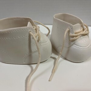 May include: A pair of off-white ceramic baby shoes featuring a smooth matte finish, molded stitch detailing along the sides, a rounded toe cap with perforated accents, and thin, light-colored fabric laces threaded through the eyelets.