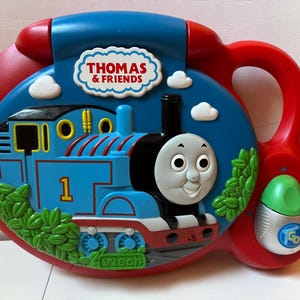 May include: A red and blue VTech Thomas & Friends electronic learning toy featuring a blue train with the number 1, a smiling face, and green bushes. The toy includes a white cloud-shaped logo, a handle, and a green button labeled GO.