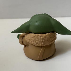 May include: A small figurine of a character with large green ears and a tan coat with a furry collar. The figure is sitting down, with its back to the viewer. The figurine is a collectible item.