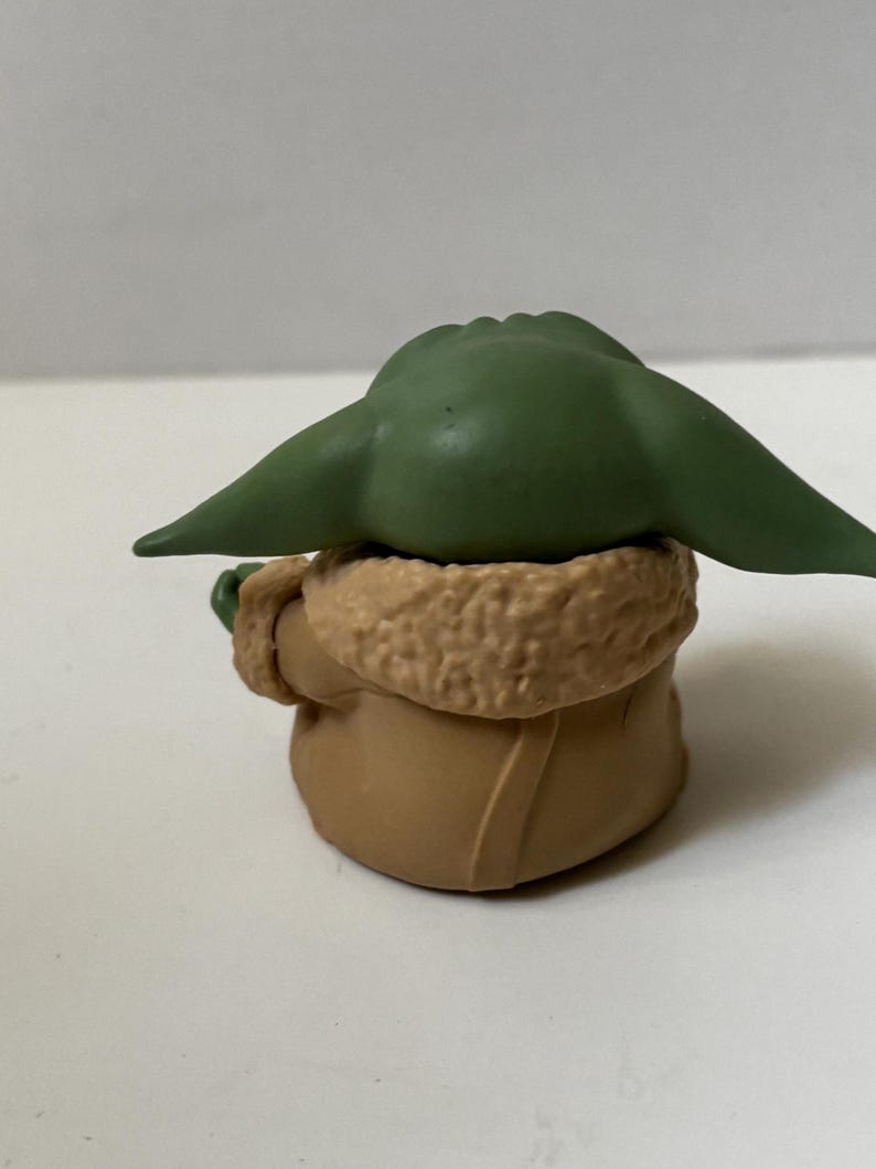 May include: A small figurine of a character with large green ears and a tan coat with a furry collar. The figure is sitting down, with its back to the viewer. The figurine is a collectible item.