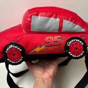May include: A red plush backpack shaped like a race car, featuring the number 95 in black and yellow, a yellow lightning bolt graphic, and black wheels with the text Lightyear printed in white. The bag includes black adjustable shoulder straps.