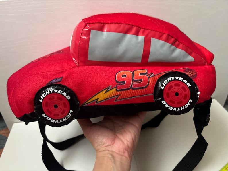 May include: A red plush backpack shaped like a race car, featuring the number 95 in black and yellow, a yellow lightning bolt graphic, and black wheels with the text Lightyear printed in white. The bag includes black adjustable shoulder straps.