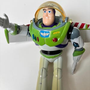 以下が含まれることがあります： クラシックなポーズのバズ・ライトイヤーのアクションフィギュア。おもちゃは、紫色のアクセントが付いた緑と白の宇宙服、透明なヘルメット、笑顔の顔が特徴です。翼は赤と白のストライプです。