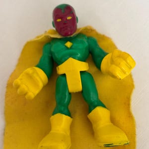 May include: A Vision action figure with a green body, yellow gloves and boots, and a yellow cape. The figure has a red face and a yellow diamond on its chest. The toy is a superhero collectible.
