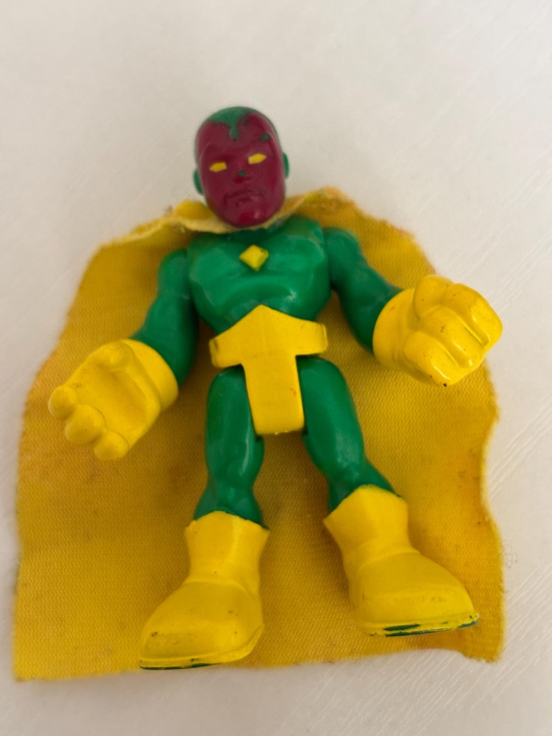 May include: A Vision action figure with a green body, yellow gloves and boots, and a yellow cape. The figure has a red face and a yellow diamond on its chest. The toy is a superhero collectible.