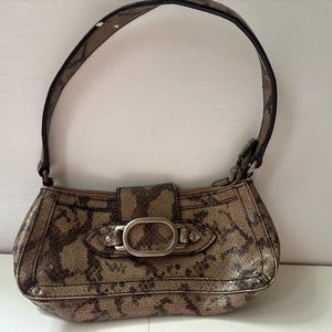 May include: A brown and tan snakeskin-patterned handbag with a curved shape and a single handle. The bag features a silver-colored buckle closure and a zipper. The handle is brown with a snakeskin pattern. The bag is designed to be carried on the shoulder.