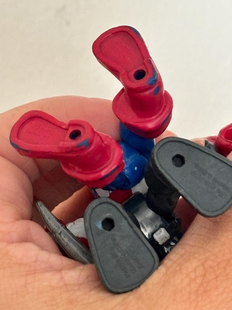 May include: A close-up of a toy action figure's legs and feet. The figure has red boots, blue pants, and black feet. The figure is lying on its back, with the feet pointing upwards.